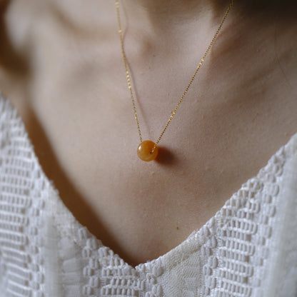 A delicate gold chain necklace with a smooth, round amber-orange bead pendant, worn on a model's neck over a white textured lace top.
